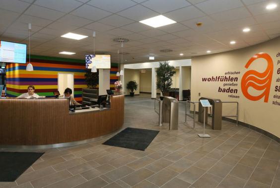 Empfangsbereich mit rundem Holztresen, zwei Mitarbeitern und zwei Monitoren, bunte Wand im Hintergrund, Wand mit orangefarbenem Logo und deutschen Worten wie 'entspannen' und 'saunieren'