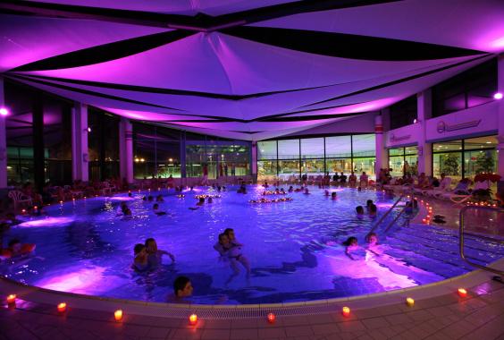 Innenpool mit vielen Menschen, beleuchtet in violettem Licht, umgeben von Kerzen und weißen Liegestühlen