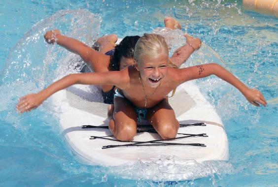 Zwei Kinder rutschen lachend auf einem weißen Surfbrett im Schwimmbecken, Wasser spritzt um sie herum