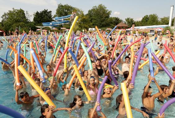 Viele Menschen unterschiedlichen Alters im Schwimmbecken eines Freibads halten bunte Schwimmnudeln in die Luft, im Hintergrund eine blaue Wasserrutsche und Bäume