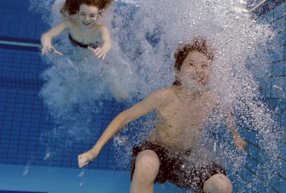 Zwei Kinder tauchen unter Wasser in einem Schwimmbecken mit blauen Kacheln und schwarzen Linien auf dem Beckenboden