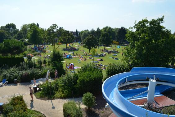 Blick auf eine grüne Liegewiese mit vielen Menschen und Sonnenschirmen, rechts eine blaue Wasserrutsche, umgeben von Bäumen und Sträuchern