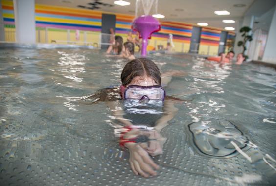 Kind mit Schwimmbrille schwimmt in einem Hallenbad, im Hintergrund weitere Kinder und bunte Wände