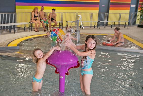 Zwei Kinder spielen an einem lila Wasserspiel im flachen Schwimmbecken eines Hallenbads, im Hintergrund sitzen drei Erwachsene auf einer Bank und weitere Kinder spielen im Wasser.
