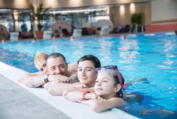 Vier Personen unterschiedlichen Alters lehnen entspannt am Beckenrand eines Hallenbads, zwei Kinder tragen Schwimmbrillen