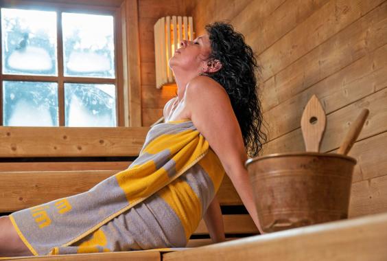 Frau mit dunklen lockigen Haaren entspannt in einer Sauna, trägt ein gelb-grau gestreiftes Handtuch, sitzt auf Holzbänken neben einem Holzeimer mit Kelle