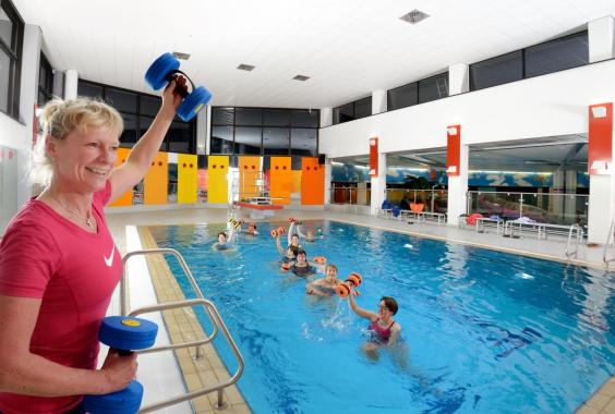 Frau in rotem Shirt hält blaue Hanteln am Beckenrand eines Hallenbads, mehrere Personen machen Aquagymnastik mit orangen Hanteln im Wasser
