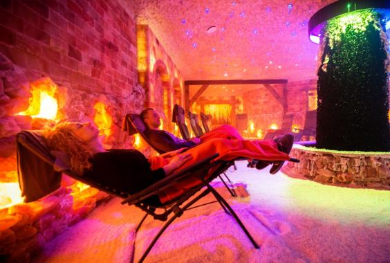 Zwei Personen entspannen auf Liegestühlen in einem warm beleuchteten Raum mit Kamin und einem beleuchteten Wasserfall