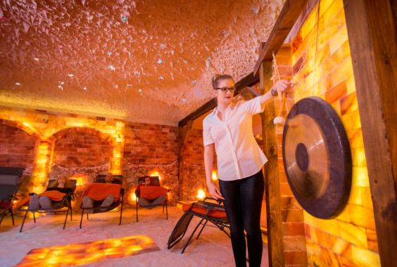 Frau schlägt Gong in einem warm beleuchteten Salzraum, während drei Personen auf Liegestühlen mit orangefarbenen Decken entspannen