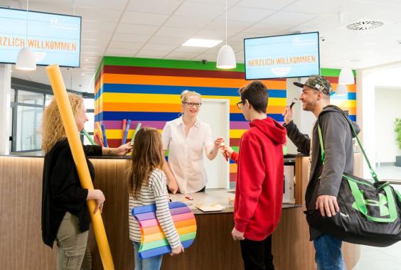 Empfangsbereich mit bunter Wand, Empfangsdame begrüßt eine Familie mit zwei Kindern und einem Mann mit Sporttasche