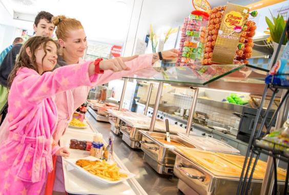 Drei Personen in Bademänteln wählen Essen an einem Buffet mit warmen Speisen und Süßigkeiten im Hintergrund