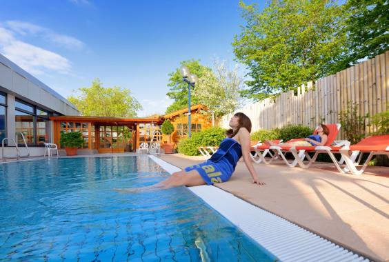 Frau mit blauem Handtuch sitzt am Rand eines Außenbeckens, andere Frau entspannt auf Liegestuhl, Sauna und Bäume im Hintergrund